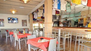 A restaurant with tables covered in red tablecloths and white chairs, a bar area with a counter, and various hanging items. - San Francisco Apartments (Sant Antoni de Portmany)
