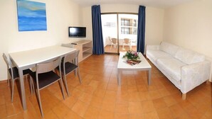 A compact living space with a dining table, chairs, a sofa, a TV, and a sliding glass door leading to an outdoor area. - San Francisco Apartments (Sant Antoni de Portmany)