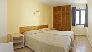 A bedroom with two beds, a nightstand, a lamp, a wardrobe, and a window with curtains. - San Francisco Apartments (Sant Antoni de Portmany)