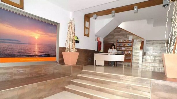 A reception area with a large painting of a sunset over the ocean, a reception desk, and a staircase leading to upper floors. - San Francisco Apartments (Sant Antoni de Portmany)