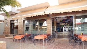 An outdoor dining area with tables and chairs, a tiled floor, and a building entrance with glass doors. - San Francisco Apartments (Sant Antoni de Portmany)