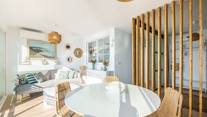 Een moderne woonkamer met een ronde tafel, rieten stoelen, een bank met gekleurde kussens, een groot raam met uitzicht, en een decoratieve wand met spiegels en een schilderij.
