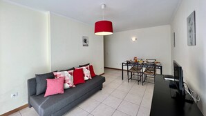 Una sala de estar moderna con un sofá gris, cojines rojos y blancos, una mesa de comedor con sillas, un televisor sobre un mueble negro y una lámpara colgante roja.