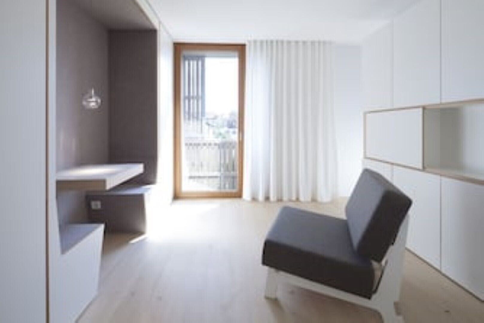 Una habitación moderna y minimalista con un banco integrado, una puerta corredera que da a un balcón y un ventanal con cortinas blancas.