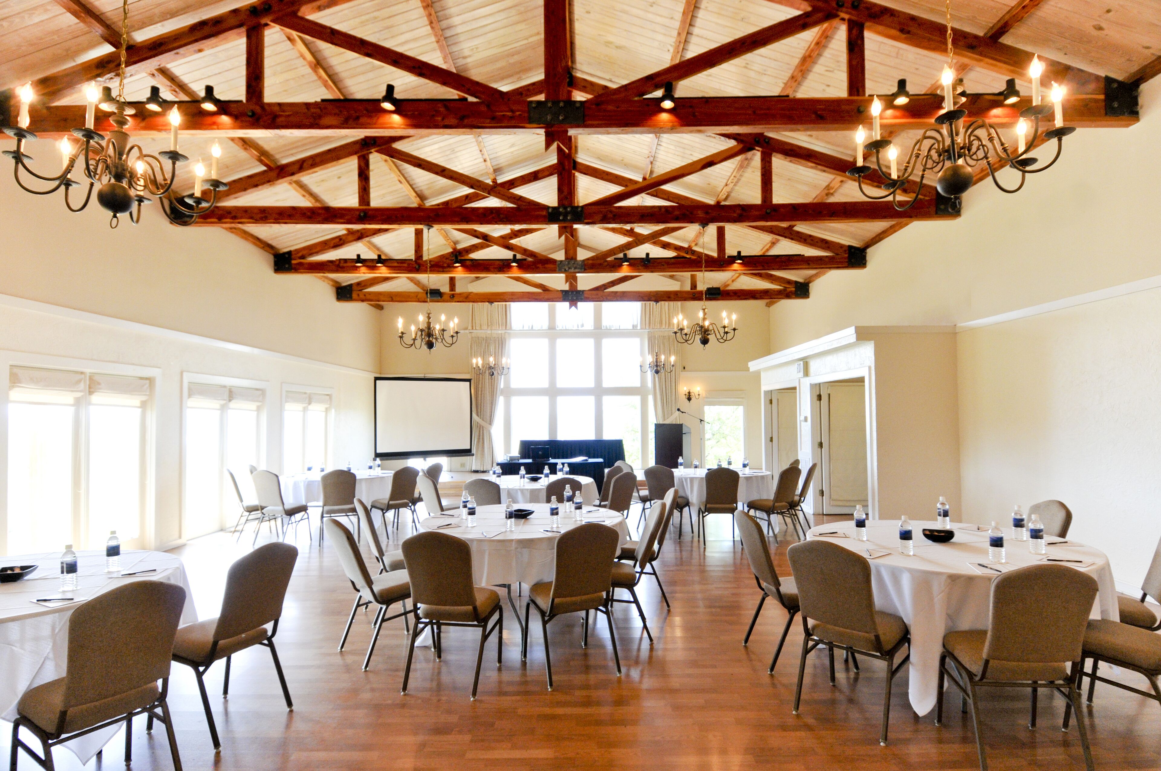 Una amplia sala de conferencias con techos altos, vigas de madera y candelabros. Mesas dispuestas para reuniones con sillas y botellas de agua.