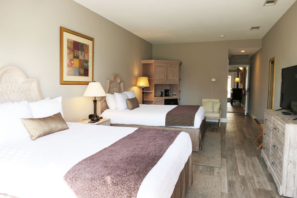A hotel room with two beds, a desk, a chair, a dresser, a television, and a hallway leading to another room.