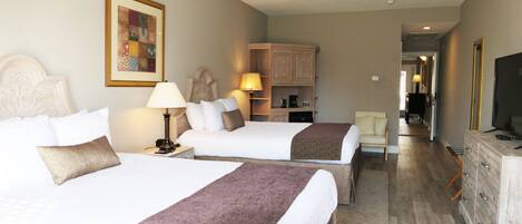 A hotel room with two beds, a desk, a chair, a dresser, a television, and a hallway leading to another room.
