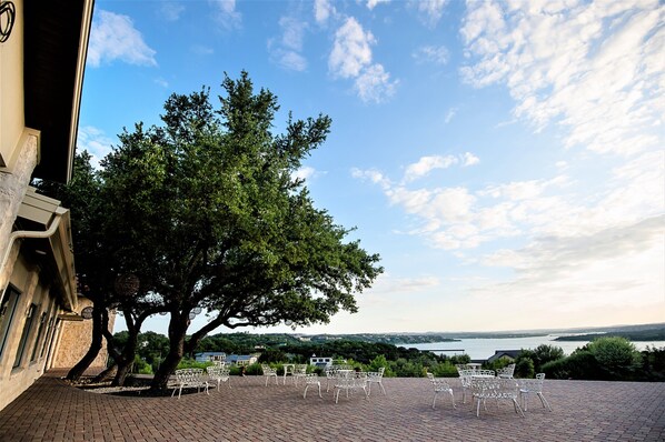 A terrace with white metal chairs and a large tree providing shade, overlooking a body of water and distant landscape.