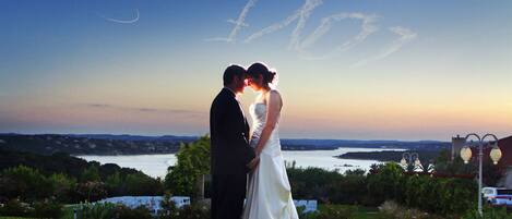 A couple standing on a grassy hill overlooking a body of water with a sunset in the background.