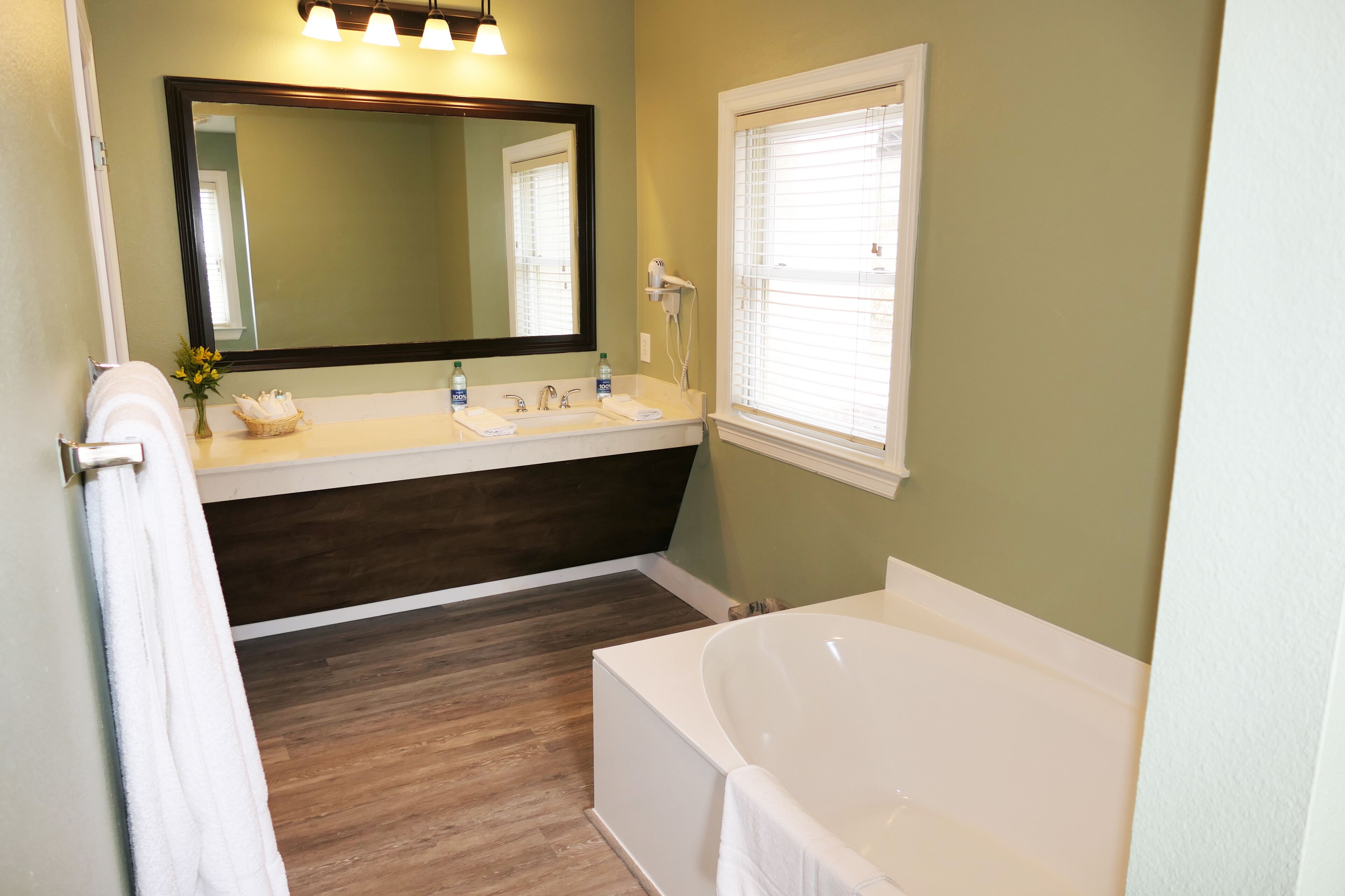 A bathroom with a bathtub, a double sink vanity, a large mirror, and a window with blinds.