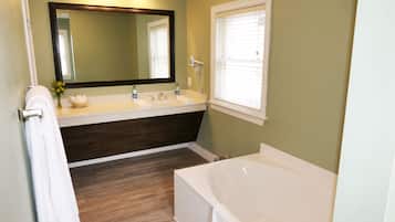 A bathroom with a bathtub, a double sink vanity, a large mirror, and a window with blinds.