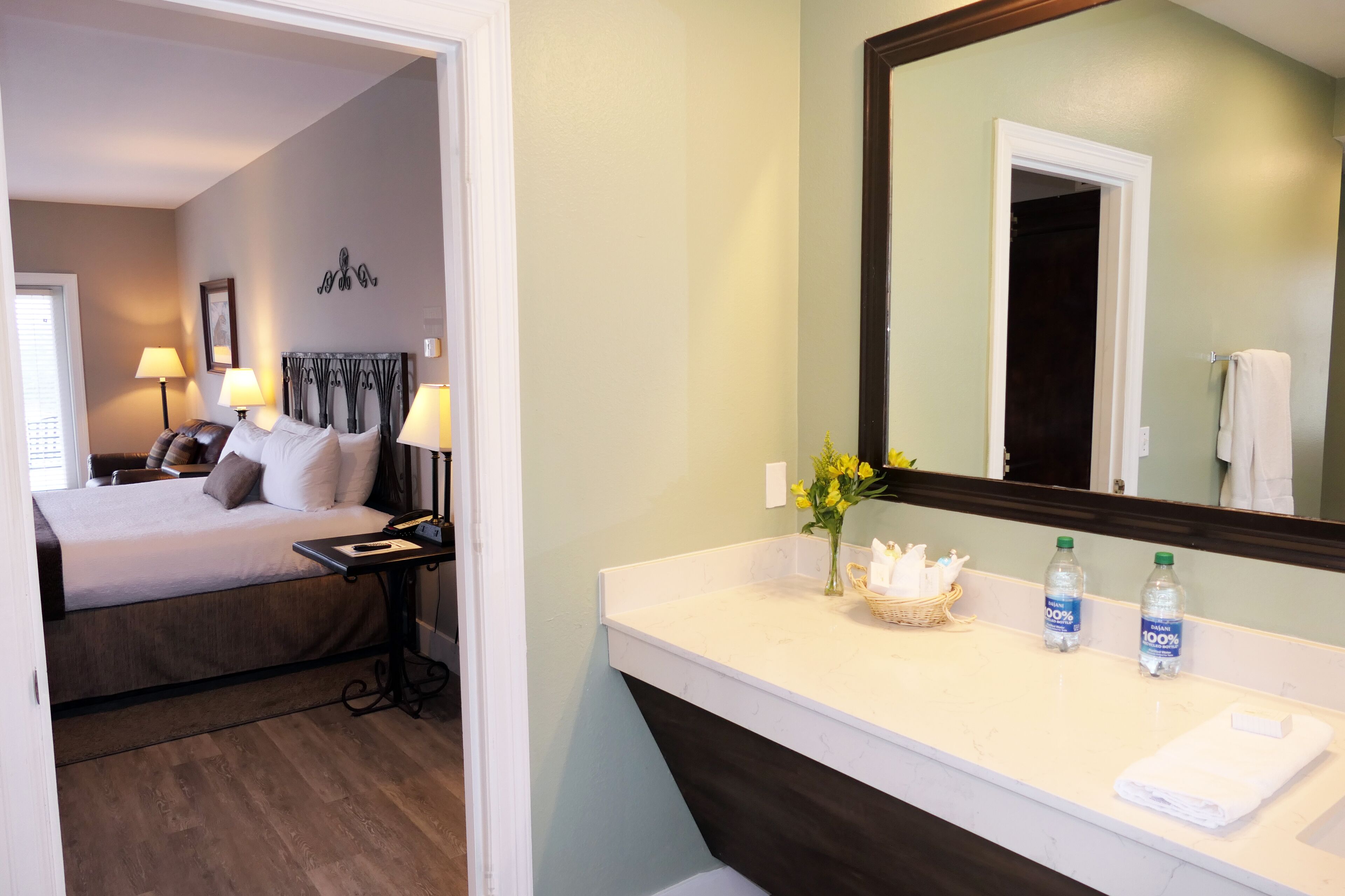 A hotel room with a bed, bedside lamps, a nightstand, a mirror, a bathroom with a sink, and toiletries.