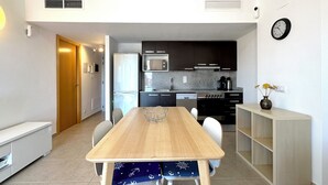 A modern kitchen with a built-in refrigerator, stove, and microwave. There is a dining table with chairs and a small shelf unit. - V019 - Mestral 1 (Roses)