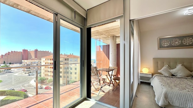 A modern bedroom with a large sliding glass door leading to a balcony, a bed with a patterned bedspread, and a wall-mounted light. - V019 - Mestral 1 (Roses)