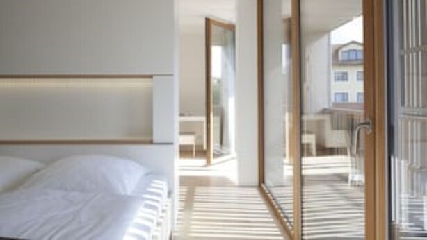 Ein modernes Hotelzimmer mit einem großen Bett, einer Glastür, die auf einen Balkon führt, und Blick auf die Stadt.