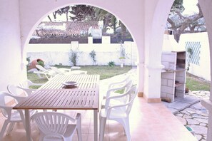 Une terrasse extérieure avec une table en bois et des chaises en plastique blanches, entourée d’un mur blanc et d’un petit jardin.