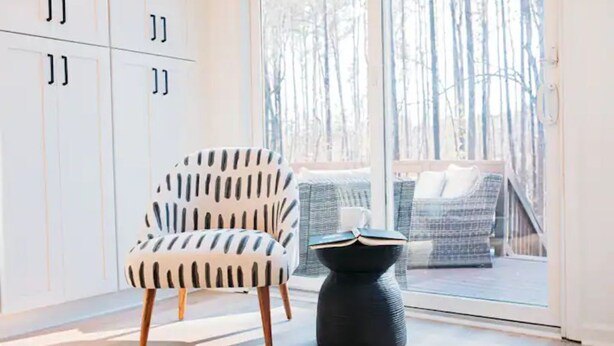 A modern room with a white chair, a black side table, and a view of a balcony with a wicker sofa. - Newly renovated modern & spacious home complete with a private pool! (Apex)
