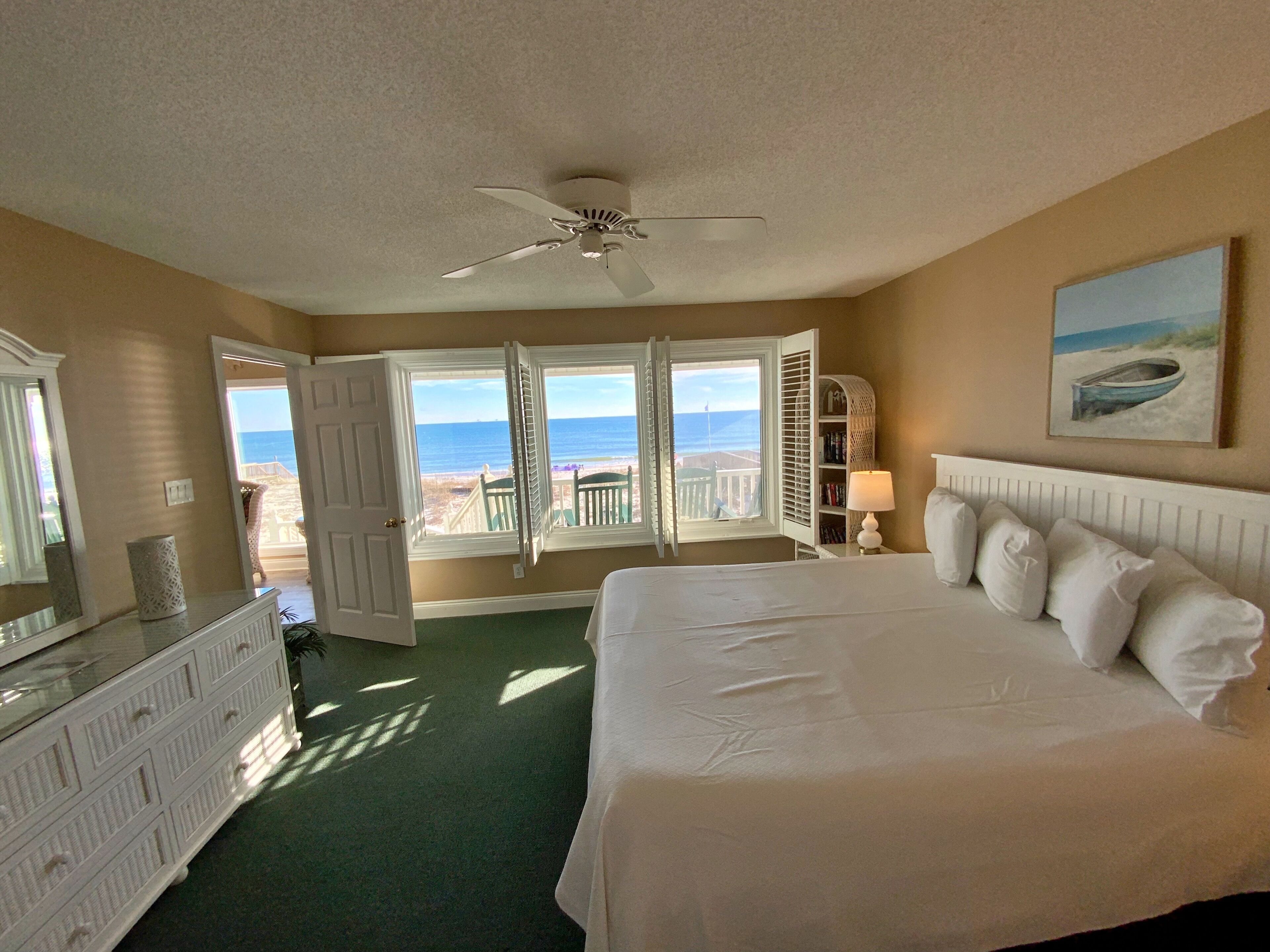 Une chambre avec un grand lit, un ventilateur de plafond, une vue sur la plage et une fenêtre avec des volets.