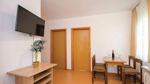 Una habitación con un escritorio de madera, un televisor en la pared, dos puertas de madera, una mesa de comedor con sillas y una planta en maceta.