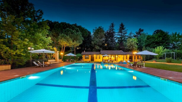 A swimming pool with a tiled deck, surrounded by outdoor seating and umbrellas, with a building in the background.