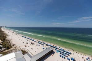 A beach with rows of blue umbrellas and sun loungers, a clear blue sky, and the ocean.