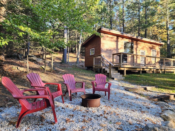 Uma cabana com varanda e escadas, rodeada por árvores. Há quatro cadeiras Adirondack e um local para fogueira.