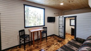 Une pièce aux murs en bois, avec une grande fenêtre, un téléviseur, une table en bois entourée de chaises et un tapis à motifs.
