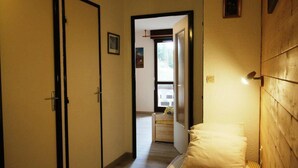Un estrecho pasillo que conduce a un dormitorio con una cama y una mesita de noche. Hay una puerta a la izquierda y una ventana a la derecha.