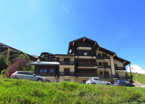 A multi-story hotel with balconies and a sloped roof, surrounded by greenery and parked cars. - 100m from resort center, 2nd floor, view mountain, balcony, tv, ski locker, 27m², Auris en Oisans (Auris)