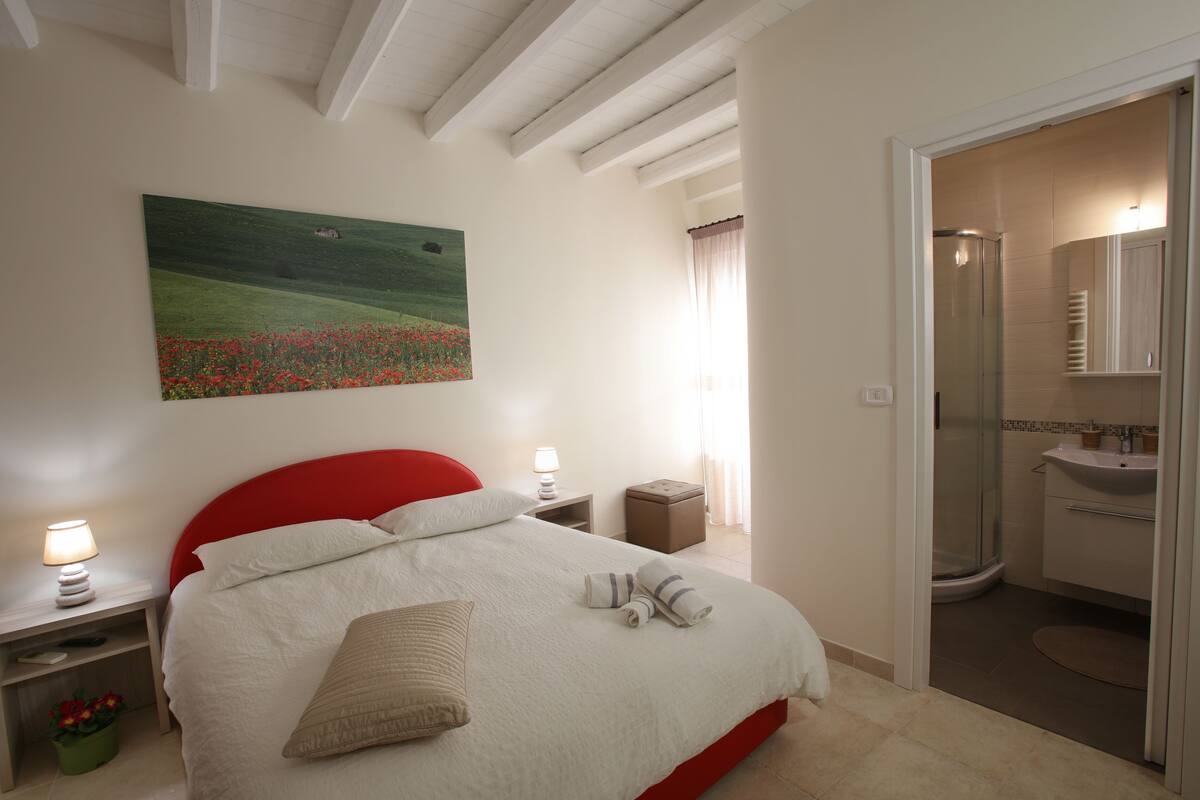 A bedroom with a red headboard, a large bed with white bedding, a painting of a field, a nightstand with a lamp, and a bathroom with a sink and shower.