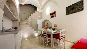A dining area with a white table, red cushioned chairs, a staircase, and a wall-mounted artwork. - CASA RUBIS (Ruvo di Puglia)
