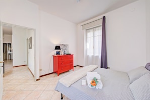 A hotel room with a bed, a red dresser, a window with curtains, and a hallway leading to another room.