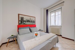 Quarto de hotel com uma cama, uma pequena mesa com uma planta, um radiador e uma janela com cortinas.