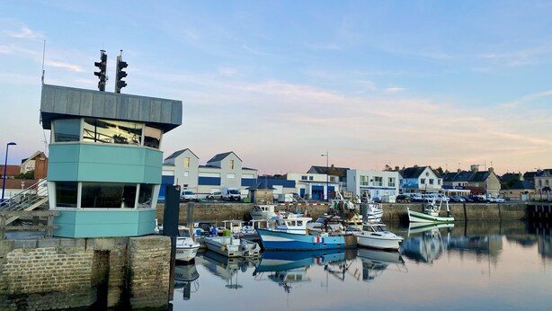 Un puerto deportivo con barcos amarrados, una torre de control y edificios al fondo.