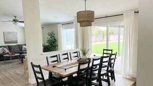 Uma sala de jantar com uma mesa de madeira e cadeiras pretas, um ventilador de teto e vista para um gramado verde através de grandes janelas.