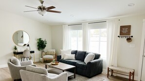 Uma sala de estar moderna com um sofá cinzento, duas poltronas, uma mesa de centro, uma escrivaninha com uma cadeira, um espelho grande e um ventilador de teto.