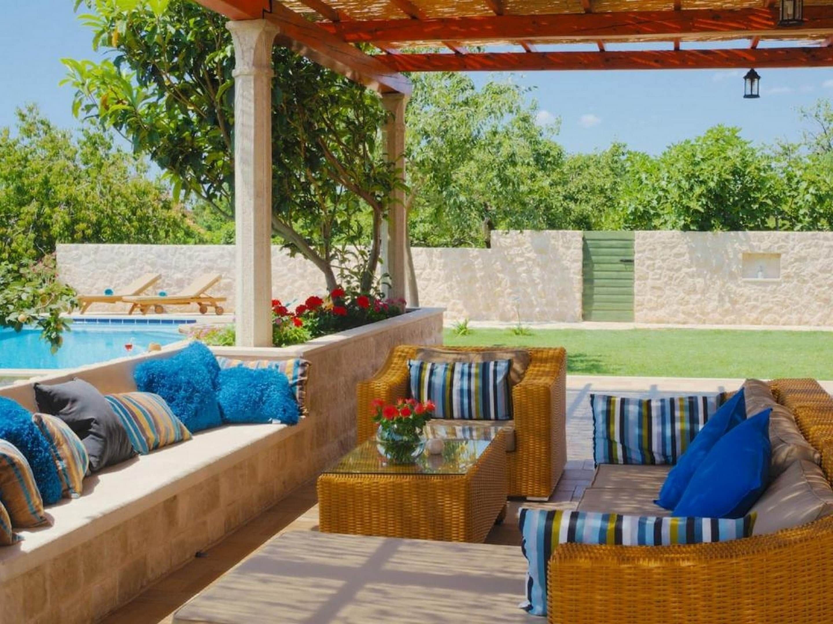 A covered outdoor seating area with wicker furniture and cushions, a pool in the background, and a stone wall.