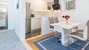 A modern kitchen with white cabinets, a stainless steel refrigerator, and a microwave. There is a dining table with two chairs and a vase of red flowers. - Apartments Lucija - Two Bedroom Apartment With Balcony and Sea View (Dubrovnik)
