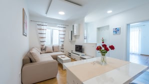 A modern living room with a sofa, coffee table, TV, and a dining area with a white table and vase of flowers. - Apartments Lucija - Two Bedroom Apartment With Balcony and Sea View (Dubrovnik)