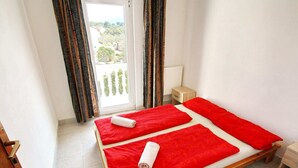 A room with a wooden bed, two red bedsheets, a white pillow, a wooden nightstand, a small drawer, and a view of greenery through a window with curtains. - Apartments Lemes - One Bedroom Apartment With Garden View Apt4 (Janjina)