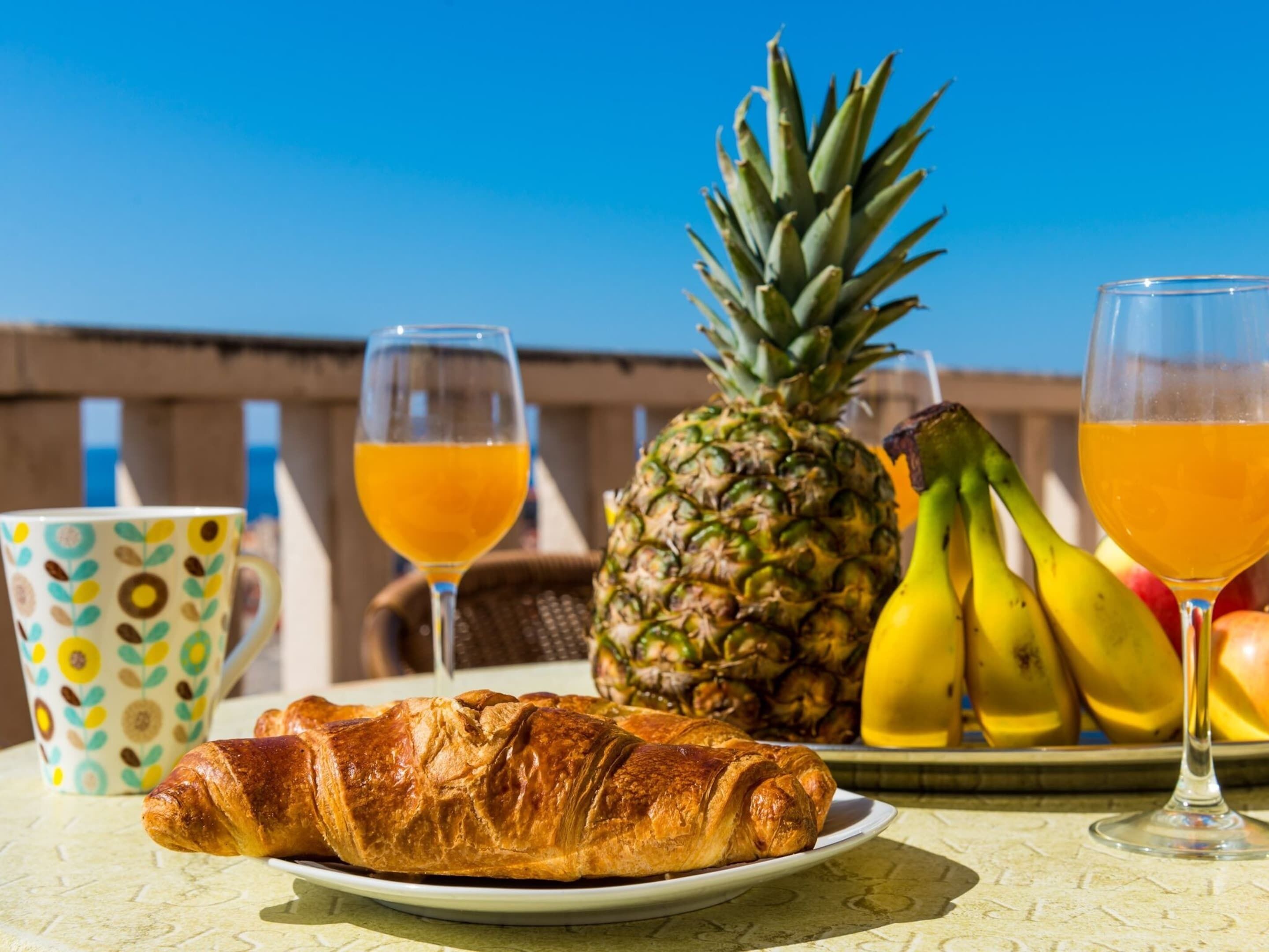 A table with a croissant, pineapple, bananas, and two glasses of orange juice.