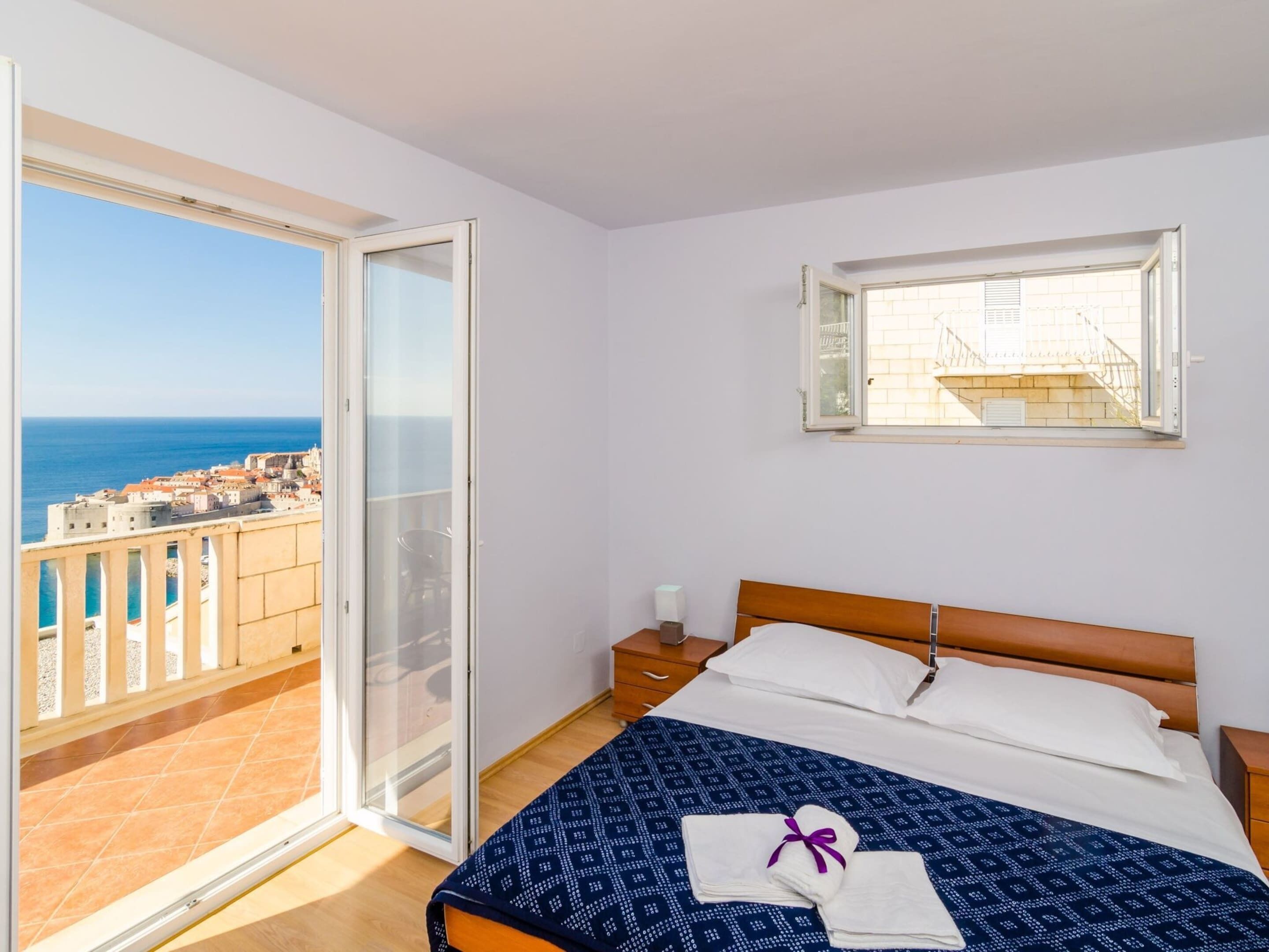 Ein Schlafzimmer mit Bett, Nachttisch, Fenster und Balkon mit Blick auf das Meer und Gebäude.