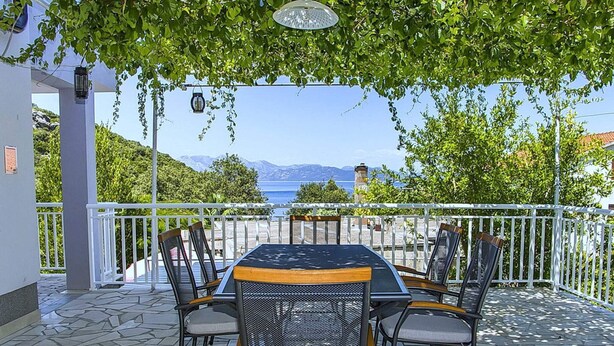 Eine Terrasse im Freien mit Tisch und Stühlen, einem weißen Geländer und Blick auf das Meer und die Berge.