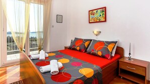 A hotel room with a bed, a nightstand, a wall-mounted lamp, a window with sheer curtains, and a view of the outdoors. - Apartments Lina - Three Bedroom Apartment With Two Balconies and Sea View (Dubrovnik)