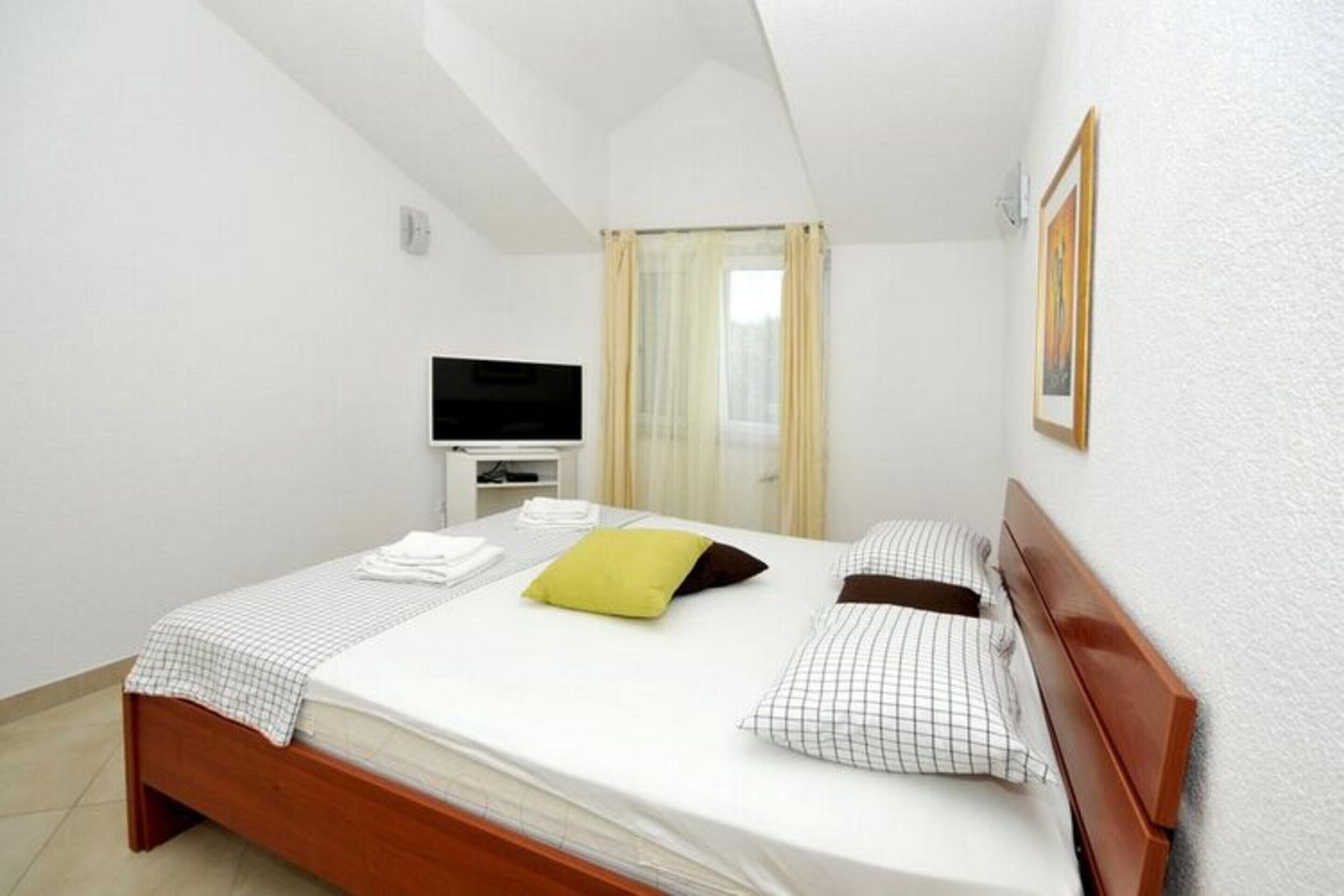 A neatly made bed with a checkered bedspread, a green pillow, and two patterned pillows. There is a television on a small white stand to the left of the bed.