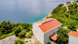 A coastal building with a red roof, surrounded by greenery and overlooking a calm sea. - Villa Kosa - Two Bedroom Apartment With Terrace and Sea View (Brela)