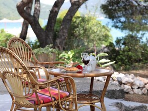 A wicker chair with a colorful cushion, a small round table with a teapot, glasses, and a croissant, set outdoors with a view of trees and a body of water. - Apartments More & Ja - One Bedroom Apartment With Terrace and Sea View (Ston)