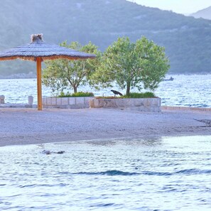 A beach with a thatched-roof structure, trees, and a bird on the shore. - Apartments More & Ja - One Bedroom Apartment With Terrace and Sea View (Ston)