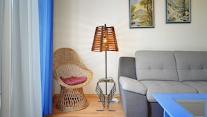 Un salon avec un fauteuil en osier, un canapé gris, une lampe sur pied et deux tableaux encadrés accrochés au mur.