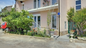 Un immeuble de deux étages doté de balcons, une voiture rouge garée devant, et diverses plantes et fleurs disposées autour de l’entrée.